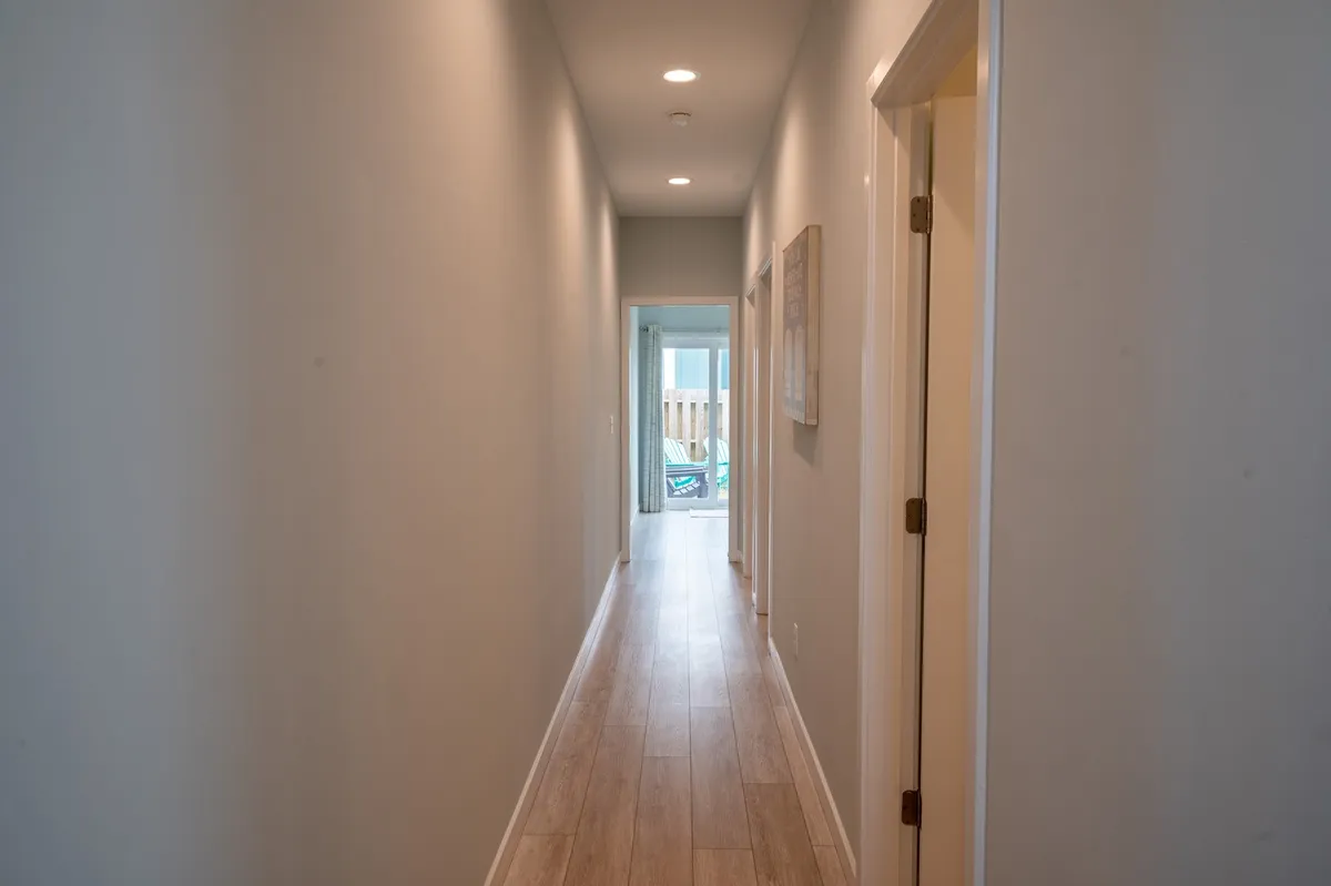 A calm, welcoming hallway leads toward a bright doorway, suggesting the transition from interior to exterior space. The view down the hallway evokes a sense of movement and exploration, hinting at journeys awaiting just outside the home.