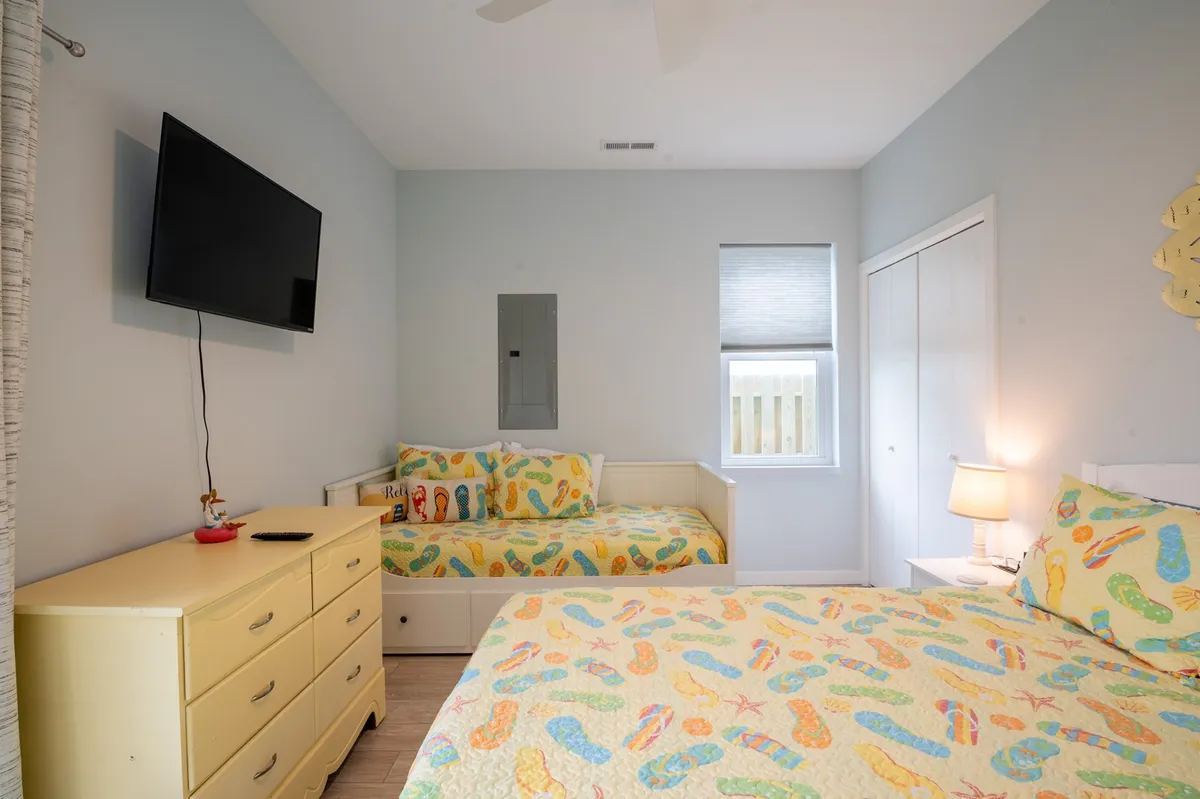 A cheerful bedroom with colorful flip-flop patterned bedding, a wall-mounted television, a light dresser, and ample natural light from the window. The playful design makes it welcoming for families or grandparents.