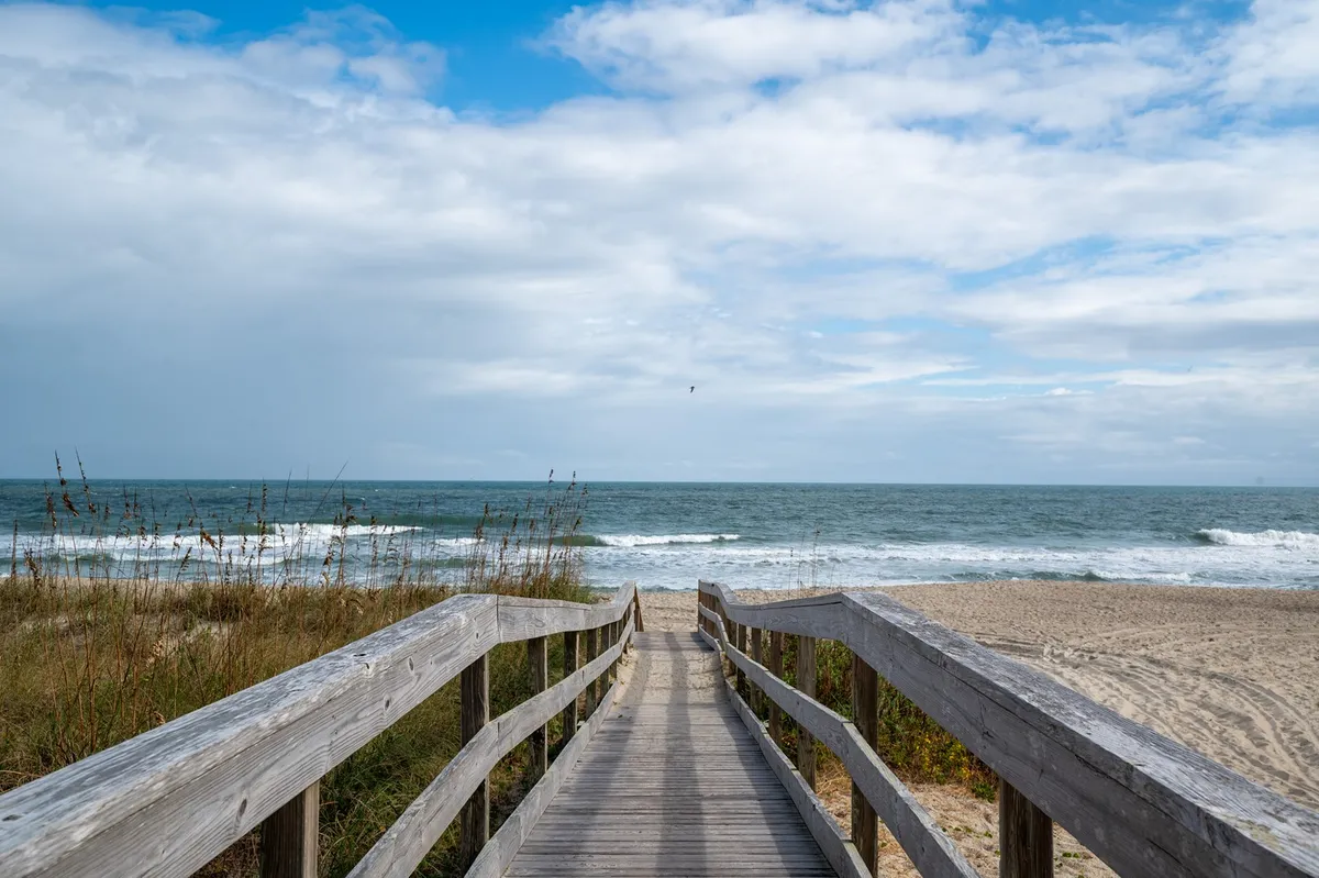 A classic wooden boardwalk leads to a sandy beach bordered by ocean waves and a serene sky. This inviting scene evokes the experience of morning strolls, easy beach access, and the tranquil natural beauty awaiting guests and pets alike.