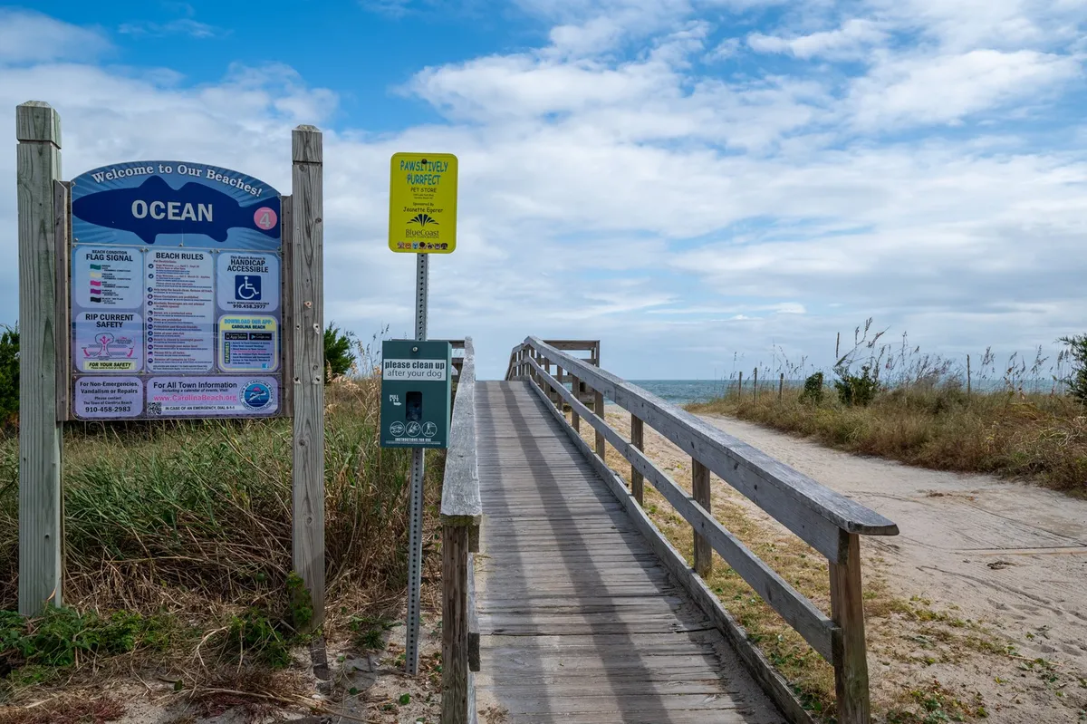 A wide wooden boardwalk leads directly to the sandy beach, with ocean views stretching into the distance. Beach access signs flank the path, inviting guests to explore the shoreline. The sky is clear, casting bright light over the scene.