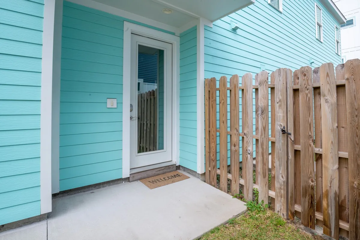 The image presents the exterior entrance to the property, with vibrant turquoise siding, a welcoming glass door, and a wooden fence beside a cheerful welcome mat. The inviting setting hints at an approachable, lively atmosphere—perfect for guests stepping out to explore local attractions or returning home after a night out.