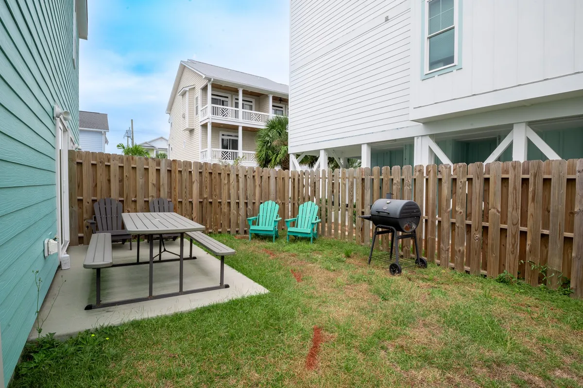 This image features a fenced backyard patio with a picnic table, outdoor chairs, a grill, and a secure fence enclosing the area. The scene exudes a casual, friendly vibe—perfect for gatherings, outdoor dining, and playtime with kids or pets. The sunlight filtering through adds a welcoming touch, and the furniture arrangement encourages socializing.