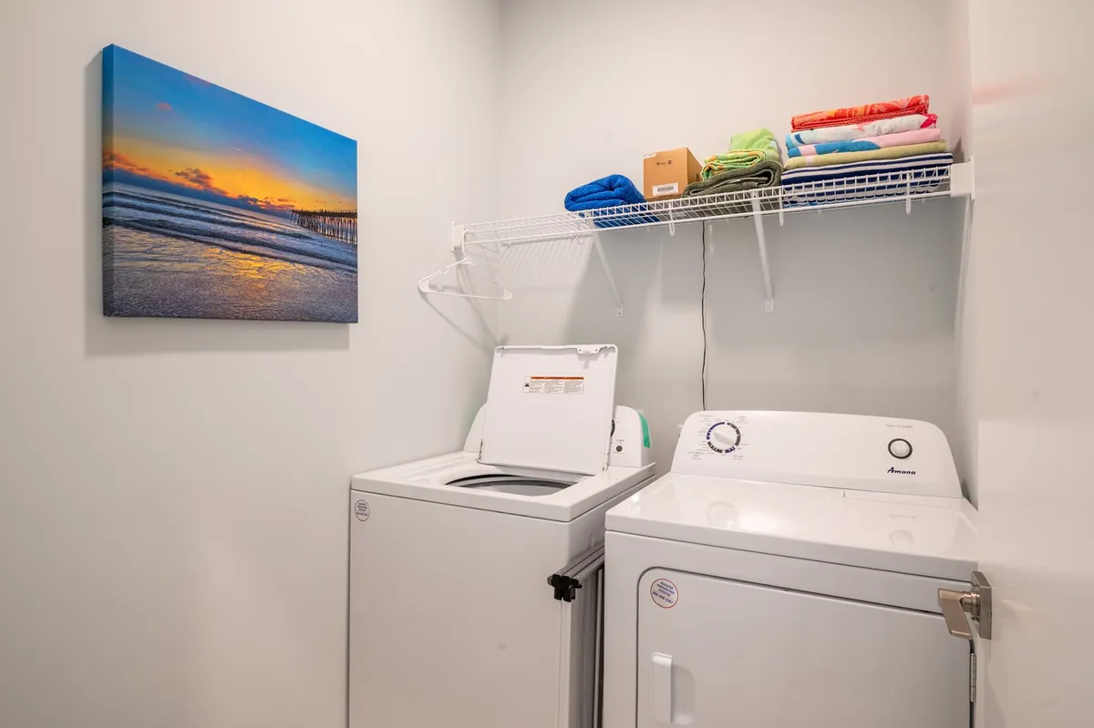 A well-appointed laundry room featuring a modern washing machine and dryer sits beside ocean-themed artwork and a shelf neatly stocked with fresh towels. The space feels organized, functional, and peaceful.